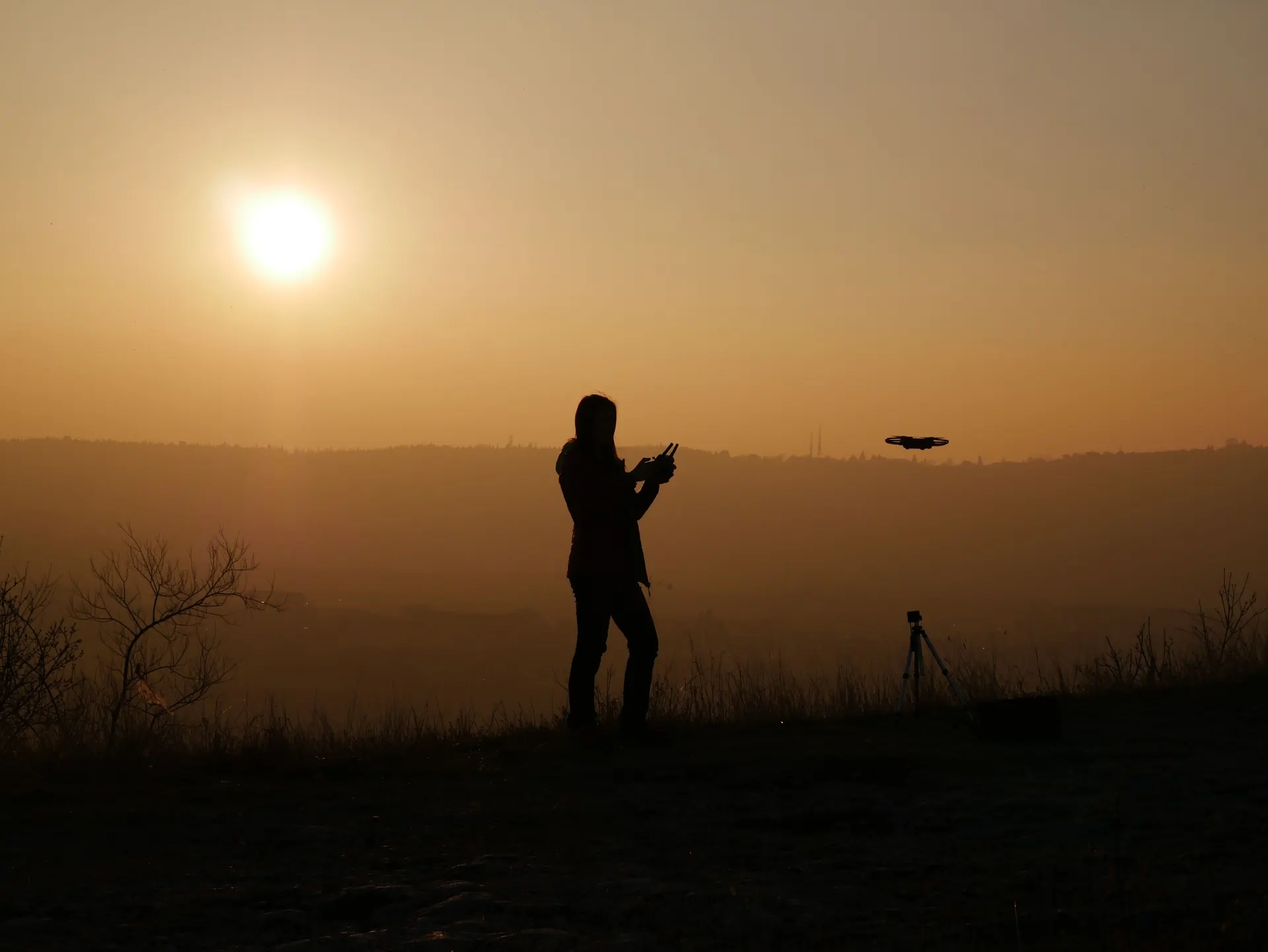Piloto com comando ao pôr do sol, com o drone em voo.